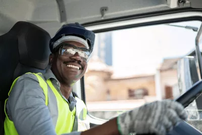 A black man wearing a safety vest, protective glasses, safety helmet and gloves while driving a truck.