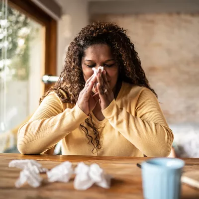 A Woman Sits at a Table with a Cup of Tea Blowing Her Nose