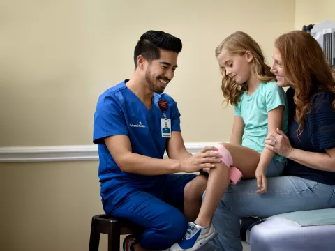 A Provider Wraps a Child Patient's Knee With Pink Gauze While a Parent Watches