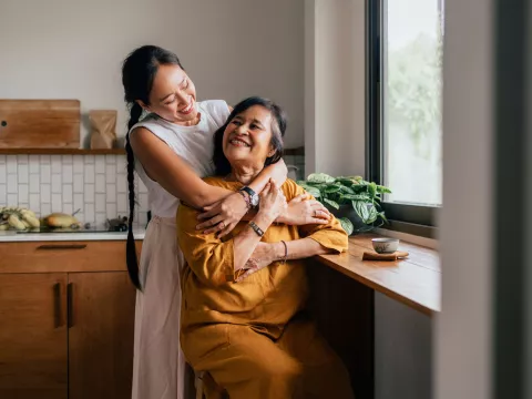 A Mother and Adult Daughter Embrace in the Kitchen in Front of a Window