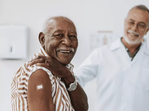 A senior man smiles and shows his arm with bandage after receiving a vaccine in the foreground while a doctor smiles in the background.