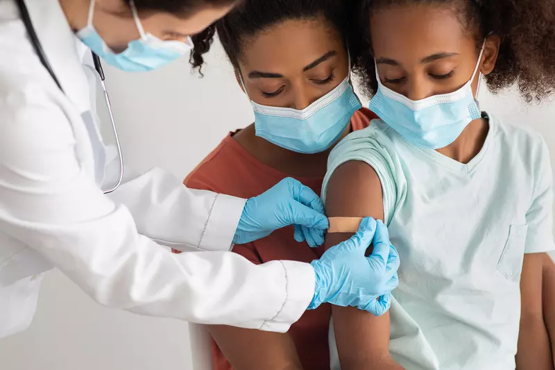 A Mother Comforts Her Daughter While a Physician Puts a Bandaid on an Injection Site in the Upper Arm. 