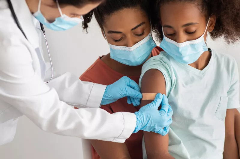 A Mother Comforts Her Daughter While a Physician Puts a Bandaid on an Injection Site in the Upper Arm.