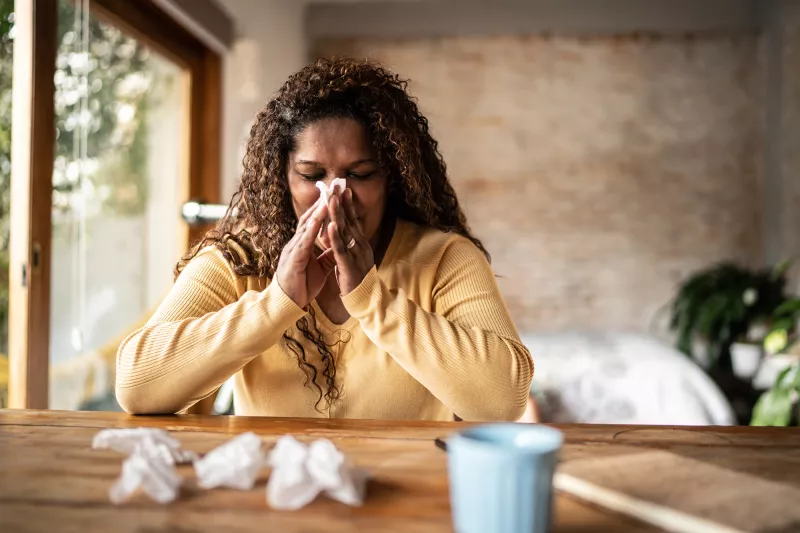 A Woman Sits at a Table with a Cup of Tea Blowing Her Nose