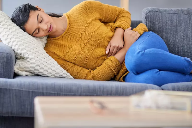 Woman, lying down on a sofa, her knees to her chest, clutching her stomach because she is experiencing endometriosis pain.