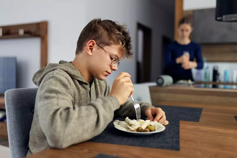 A boy sits at a dining room table eating lunch in the foreground while his mother is in the background adding seasoning to food on a plate.