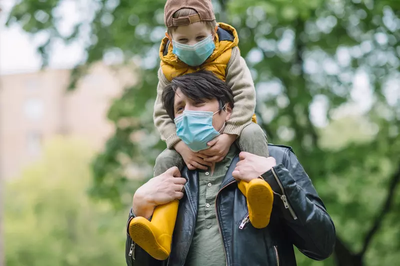 A father carrying his son on his shoulders while wearing a mask. 