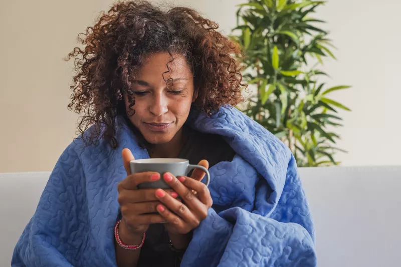 An adult black woman sips tea at home on her couch
