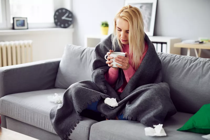 A woman drinking tea under a blanket at home.