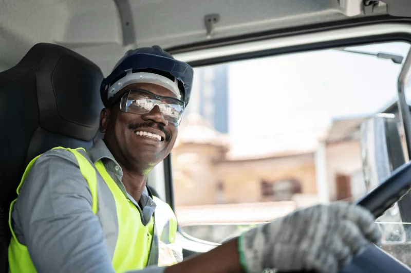 A black man wearing a safety vest, protective glasses, safety helmet and gloves while driving a truck.