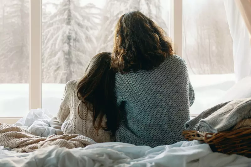 Mother and daughter looking through a window at the snow outside.