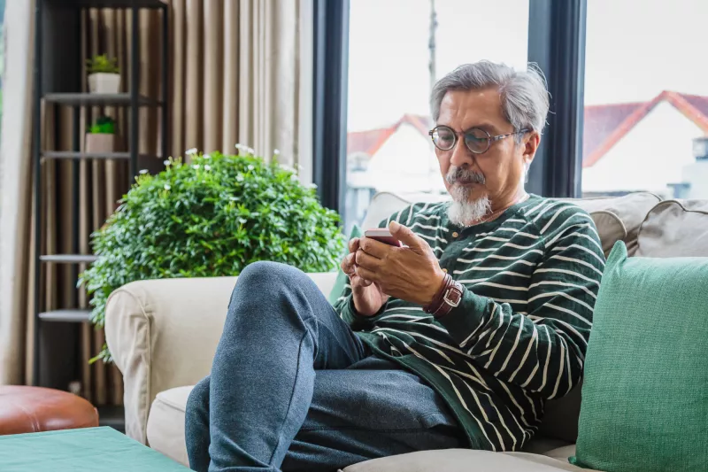 Older man sitting on a couch at home using a phone.