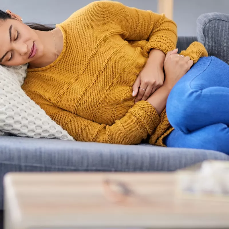 Woman, lying down on a sofa, her knees to her chest, clutching her stomach because she is experiencing endometriosis pain.