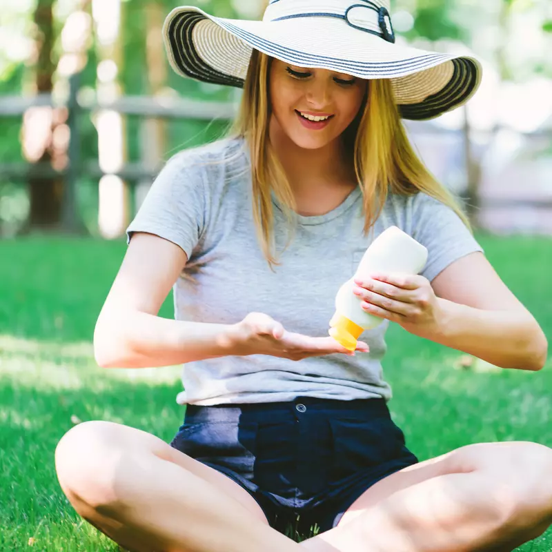 Woman, sitting on the ground wearing a hat and applying sunscreen