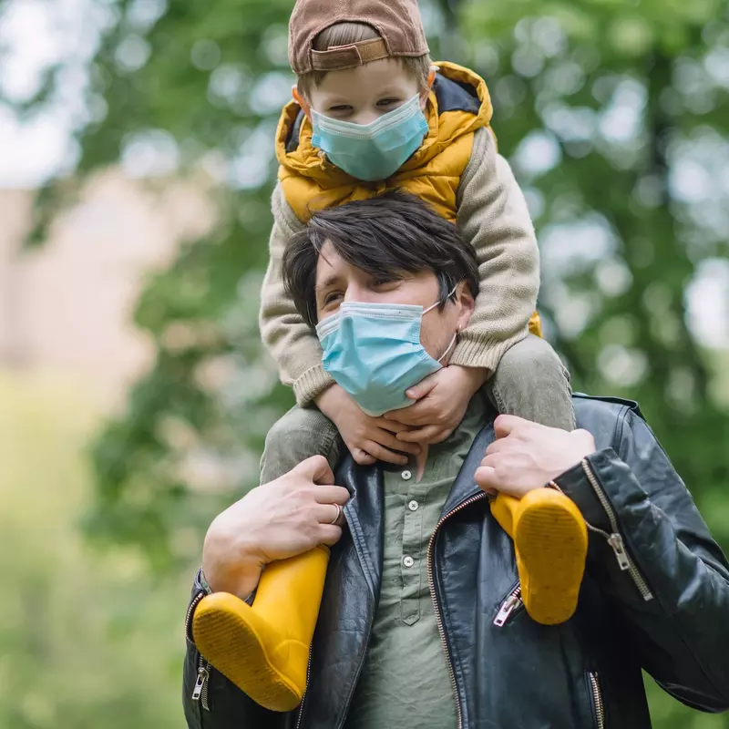 A father carrying his son on his shoulders while wearing a mask. 