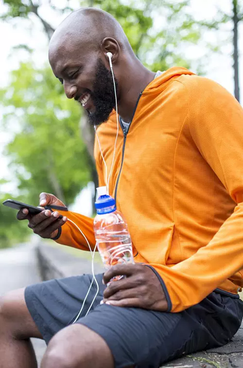 An adult man in an orange hoodie, sitting on a bench outside, looking at his phone and hydrating with a bottle of water.